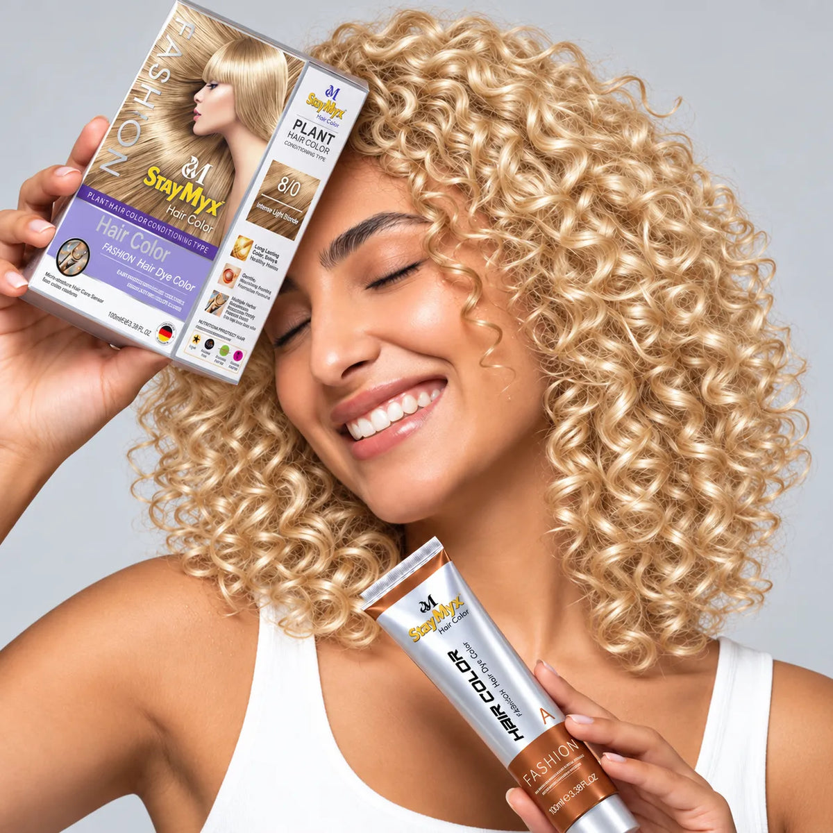 Smiling woman with voluminous, glossy blonde curly hair holding StayMyx 8/0 Intense Light Blonde hair color box and tube, showcasing vibrant blonde hair results.